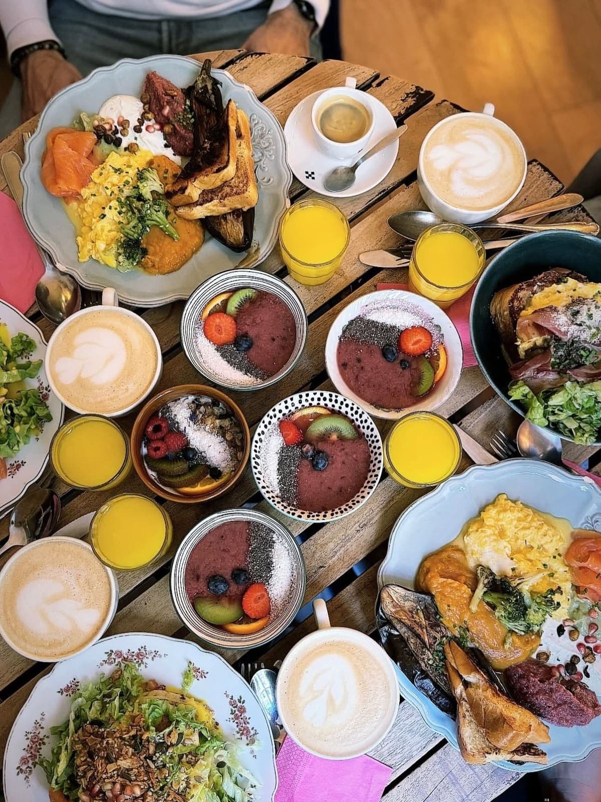 Overhead view of a brunch table with fresh orange juices, latte art coffees, smoothie bowls and savory plates