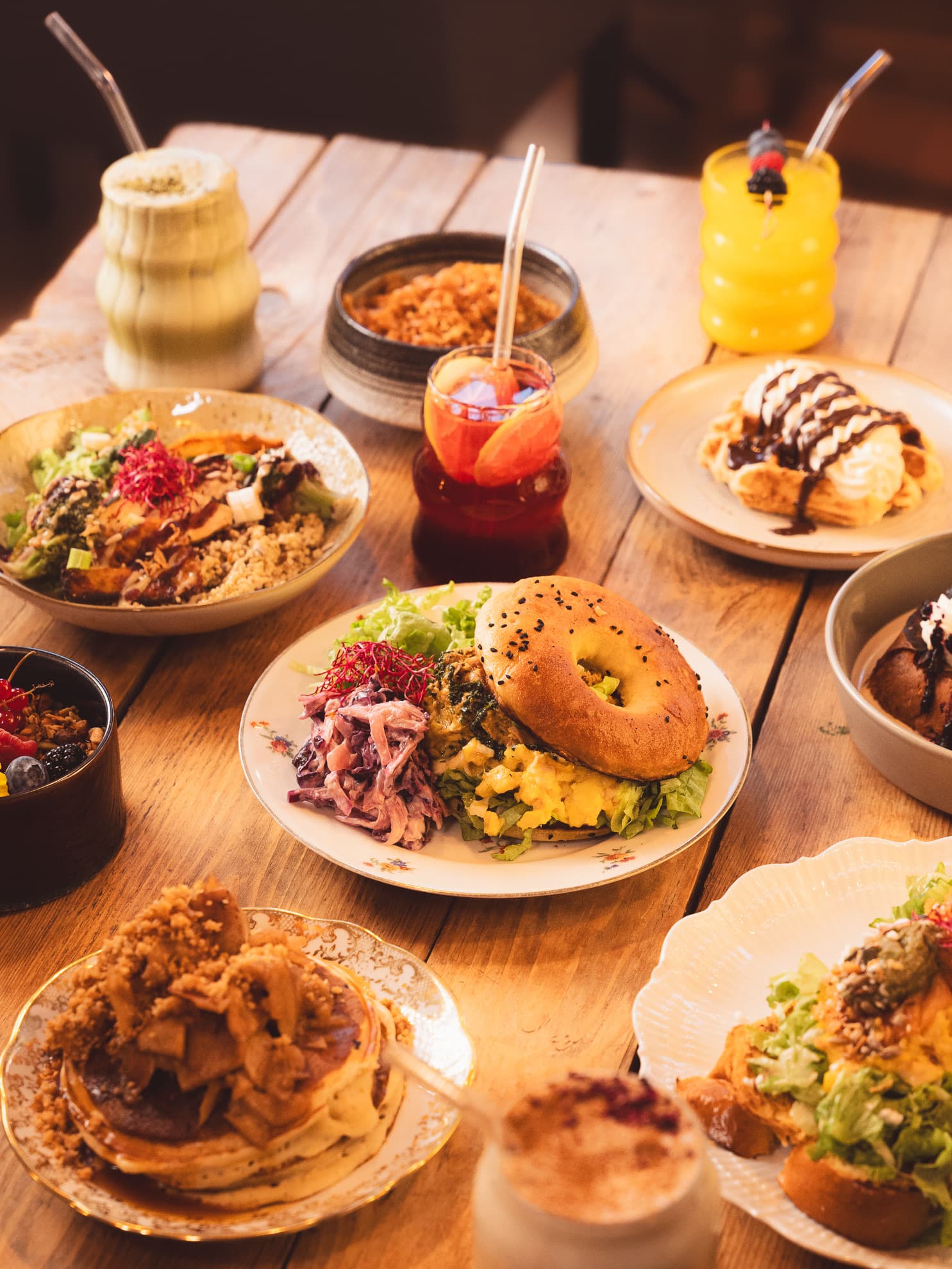 Wooden table with a bagel plate, apple crumble pancakes, a chocolate waffle, iced matcha and fresh juices