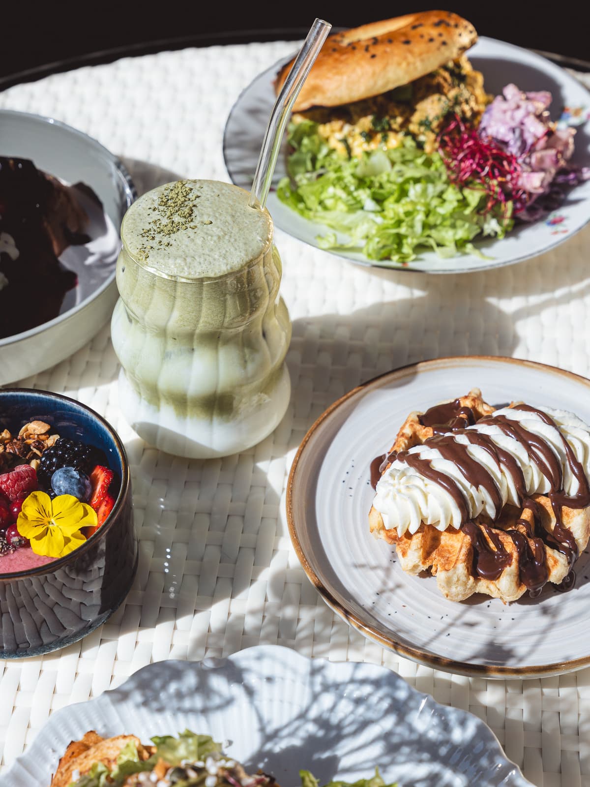 Iced matcha latte in a wavy glass beside a waffle with whipped cream and chocolate sauce