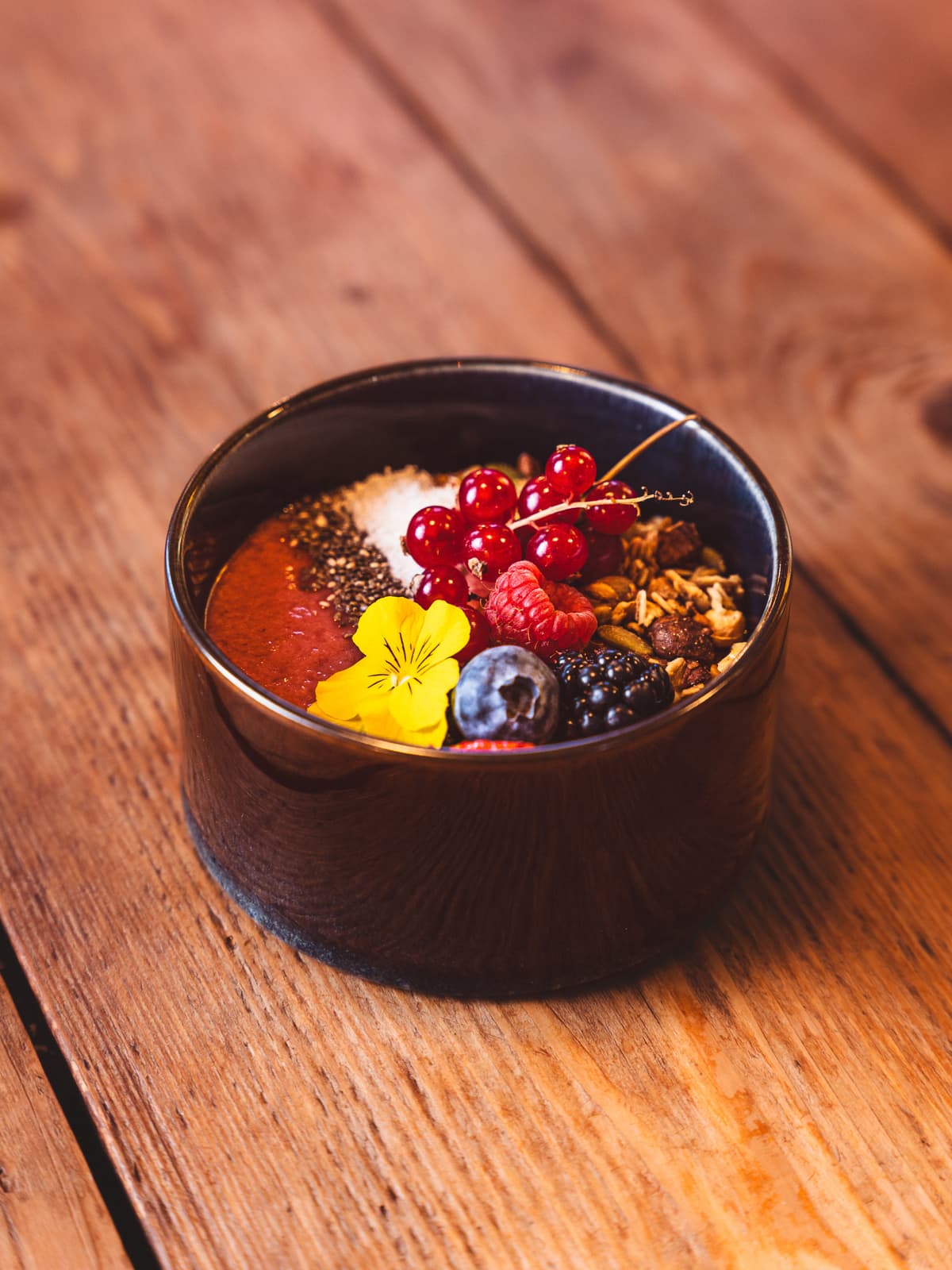 Smoothie bowl topped with homemade granola, red currants, berries, chia seeds and a yellow pansy